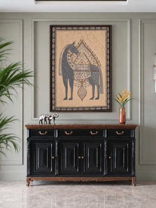 The Swarna Ashwa embroidered wall art framed above a black carved sideboard with silver elephant figures and orange blooms on a sage panelled wall.