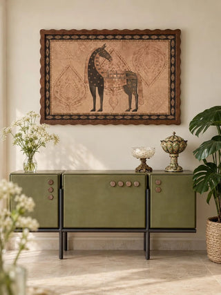 The Swarna Ashwa landscape wall art print in a scalloped walnut frame above an olive green console with white wildflowers and a crystal bowl.
