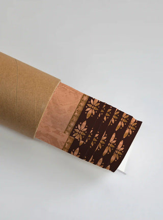 The Swarna Ashwa wall art print partly unrolled from its kraft shipping tube, showing a peach panel and a dark brown border of copper floral rosettes.