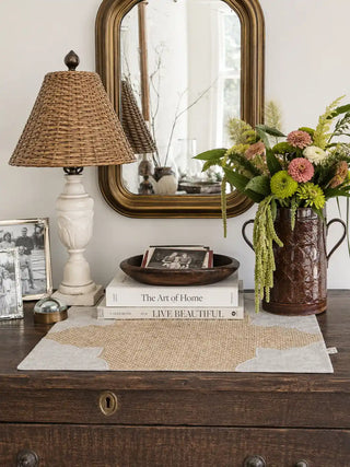 The Kasbah woven jute table mats with cream border styled on a dark wood console table alongside a rattan lamp, brass mirror, and fresh florals in a luxury home interior.