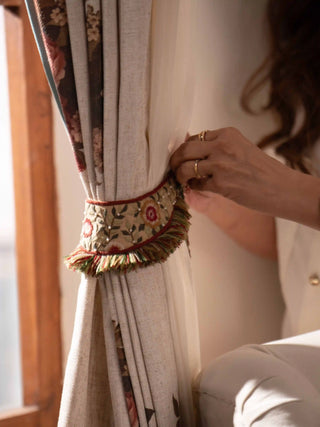 The Thiya tie back in cream with floral embroidery, pearl-like beads, red piping and a multicolour tassel fringe, gathering a curtain.