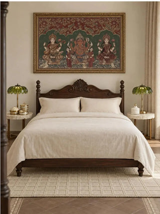 Trideva Mangalam landscape wall art in an ornate gold frame above a dark wood four-poster bed with cream linen, flanked by green leaf-shaped lamps on round side tables.