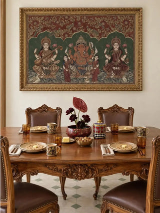 Trideva Mangalam landscape wall art in an ornate gold frame above a carved wooden dining table with leather chairs, painted mugs and a red bowl of dark flowers.