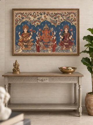 The Trideva Mangalam wall art in a gilt frame above a grey carved console with a brass figurine, a brass bowl of floating blooms and a fiddle-leaf plant.