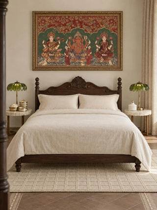 The Trideva Mangalam wall art in Rust & Forest, in an ornate gold frame, above a carved dark-wood bed with cream linen and two green palm-leaf lamps.