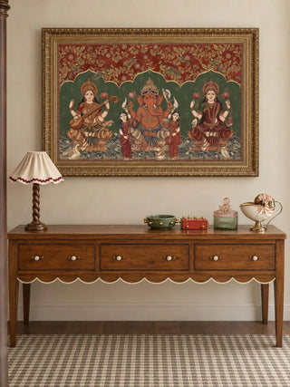 The Trideva Mangalam wall art in Rust & Forest, in an ornate gold frame, above a wooden console table with a scalloped apron, a ruffled lamp and small decorative pieces.