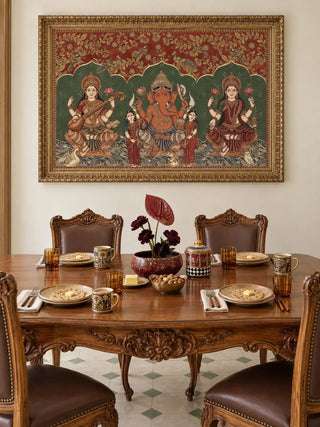 The Trideva Mangalam wall art in Rust & Forest, in an ornate gold frame, above a carved wooden dining table set with plates, mugs and dark red flowers.