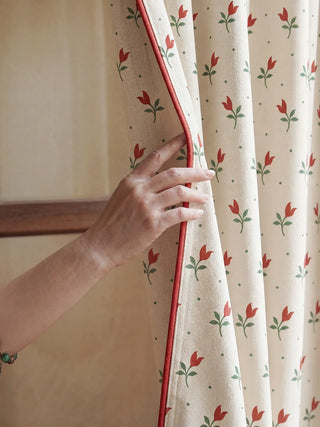 A close-up of a hand opening the Tulip Field floral curtains, featuring a sophisticated red contrast piping.