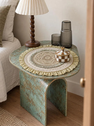 Utsavi round table mat with a gold ruffled edge and star-motif centre on a green patinated side table beside a lamp, a glass carafe and a checkered lidded box