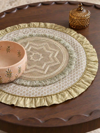 Close-up of an Utsavi table mat showing the gold ruffled edge, floral print ring, sage fringe and star-motif centre on a carved walnut table beside a peach bowl