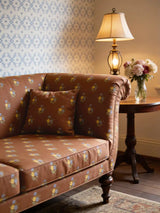 A luxury sofa and matching cushion upholstered in Vasant fabric featuring a blue and yellow floral buti pattern on chocolate brown.
