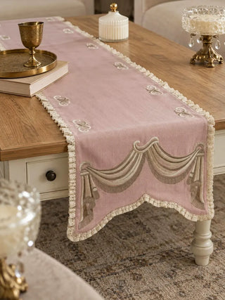 The Veloura table runner in Dusty Rose & Ecru on a wooden coffee table with a cream base, with scroll motifs and lace edging visible.