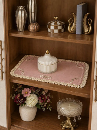 The Veloura table mat in Dusty Rose & Ecru on a wooden display shelf under a ribbed white candle jar, with brass and checkerboard accents nearby.