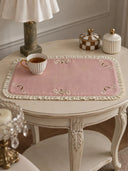 The Veloura table mat in Dusty Rose & Ecru on a round cream side table with a striped teacup and small decorative jars.