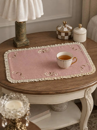 The Veloura table mat in Dusty Rose & Ecru on a round wooden-top side table with a teacup, a lamp and small decorative jars.