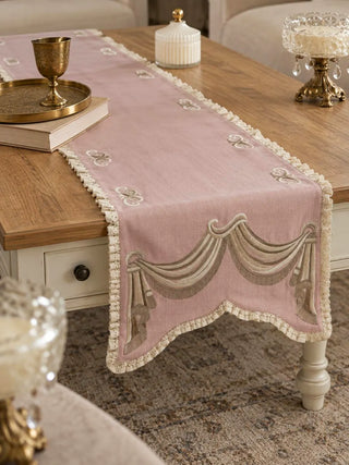 The Veloura table runner in Dusty Rose & Ecru on a wooden coffee table with a cream base, with scroll motifs and lace edging visible.
