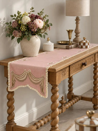 The Veloura table runner in Dusty Rose & Ecru on a carved wooden console table with barley-twist legs, styled with flowers and brass accents.