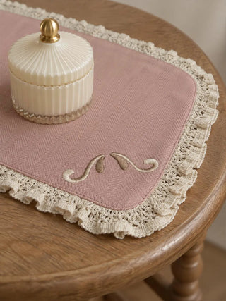 A close-up of the Veloura table mat in Dusty Rose & Ecru showing the herringbone weave, stitched scroll motif and ruffled crochet-style lace trim.