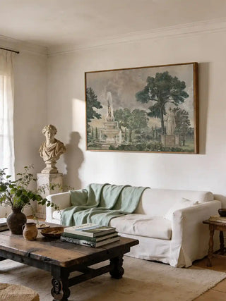 Vintage European Landscape wall art in sage green in a wooden frame above a white slipcovered sofa with a sage throw, rustic timber coffee table and classical bust in a neutral living room