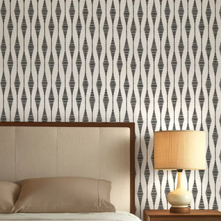 A black and white abstract wallpaper featuring a repeating wave-like geometric pattern, styled behind a bed with a lamp.