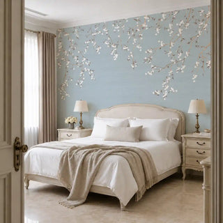 The Whispering Branches wallpaper mural in Dusty Blue installed on a feature wall in a bedroom, trailing white blossom branches above an upholstered bed frame. view-bedroom