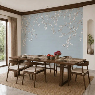 The Whispering Branches wallpaper mural in Dusty Blue installed on a feature wall in a dining room behind a wooden dining table set for a meal. view-dining