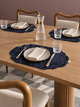 Zarfshan navy velvet placemats set at a wooden dining table with ceramic plates and ribbed glassware.