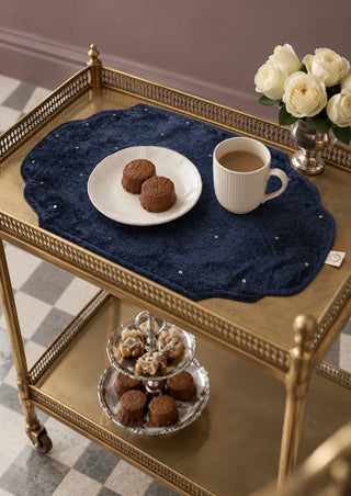 A single Zarfshan navy velvet placemat on a brass bar cart, styled with tea cakes and a cup of coffee.