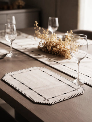 A close-up of a Zarin Trace placemat in Natural & Black beside the matching runner, styled with a wine glass and dried floral stems.