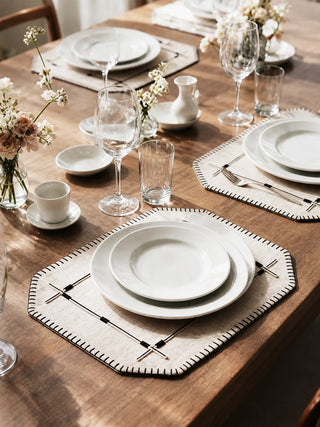 Two Zarin Trace placemats in Natural & Black set with layered ceramic plates, wine glasses and fresh flowers on a wooden dining table.
