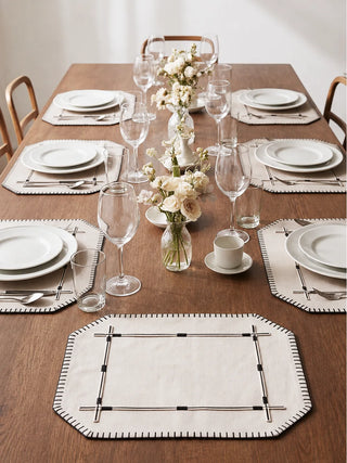Four Zarin Trace table mats in Natural & Black arranged around a set dining table with ceramic dinnerware, glassware and a floral centrepiece, viewed from above.