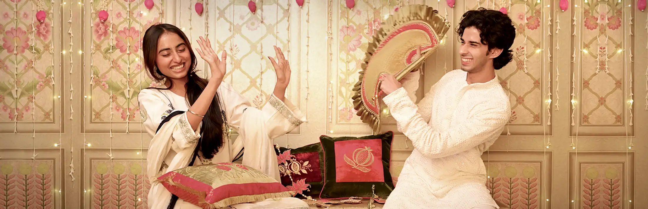 Two people in ivory festive clothes laughing with a gold hand fan and embroidered cushions against a floral panelled wall